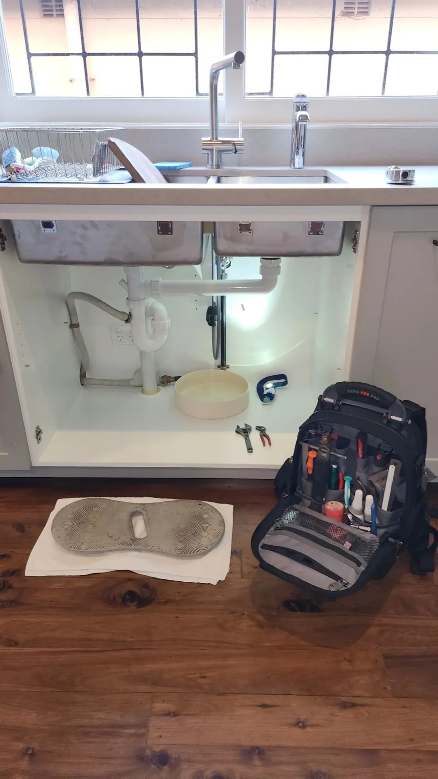Kitchen sink cupboard opened up for plumbing work, with a tool bag on the floor